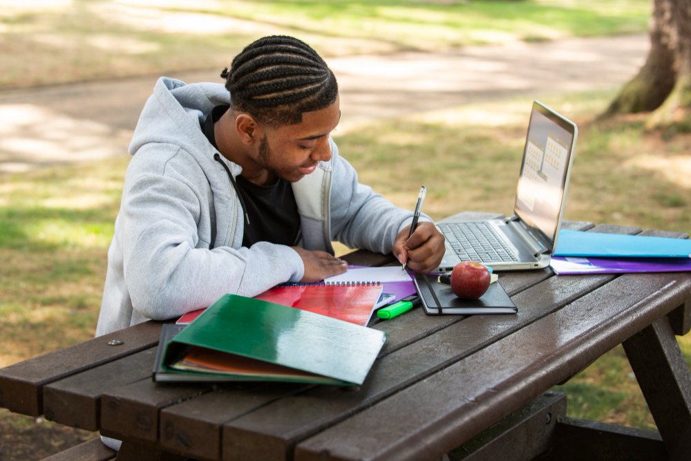 Black summer school student finishing Grade 12 math, happy teen outdoors, clear sunny path, light school supplies, academic freedom